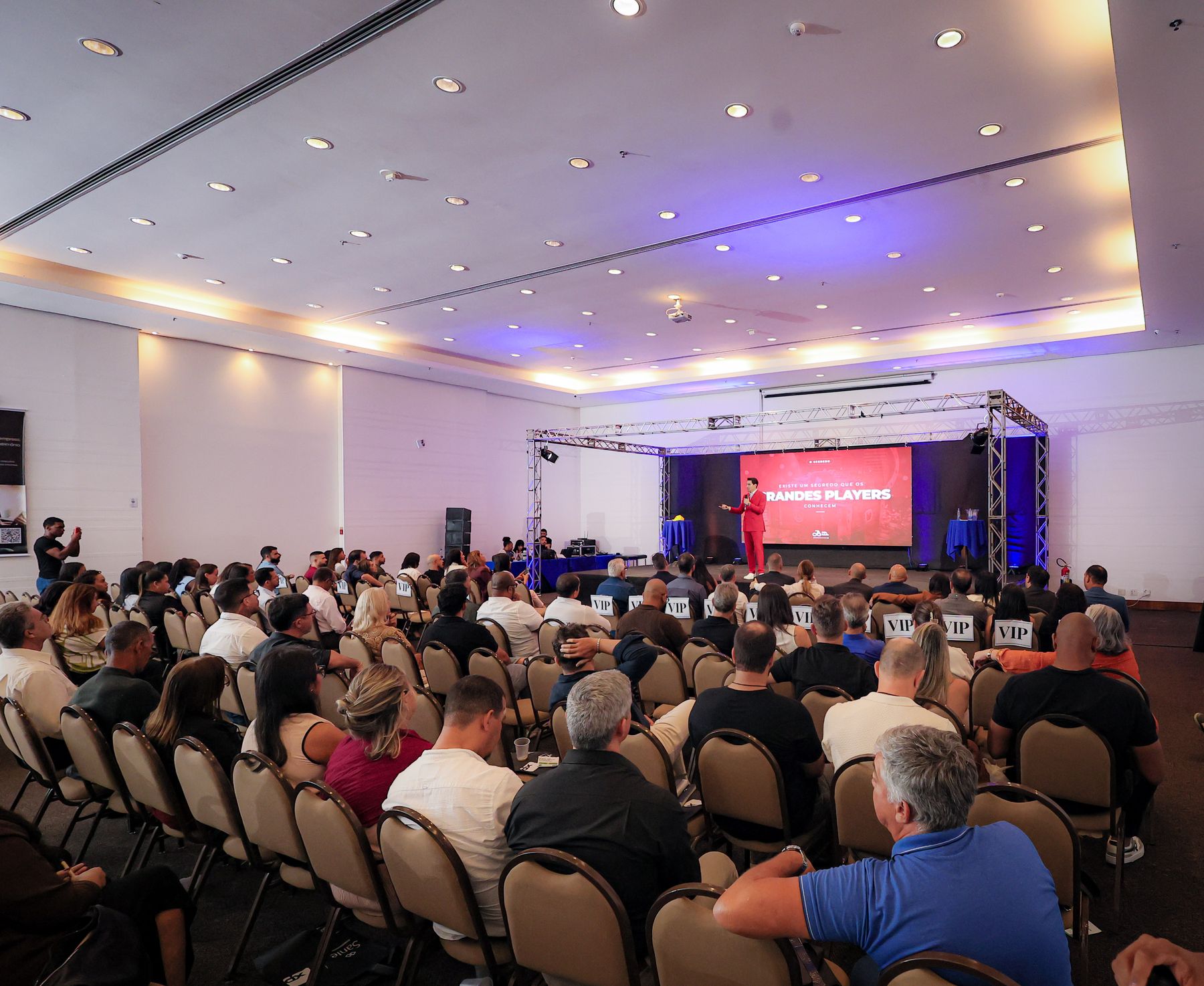 Salão do evento cheio, com a plateia voltada para o palco